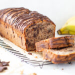Bananenkuchen aka Bananenbrot als Kastenkuchen mit aufgeschnittenen Stücken. Im Hintergrund liegen Bananen, Walnüsse und ein großes Stück Schokolade