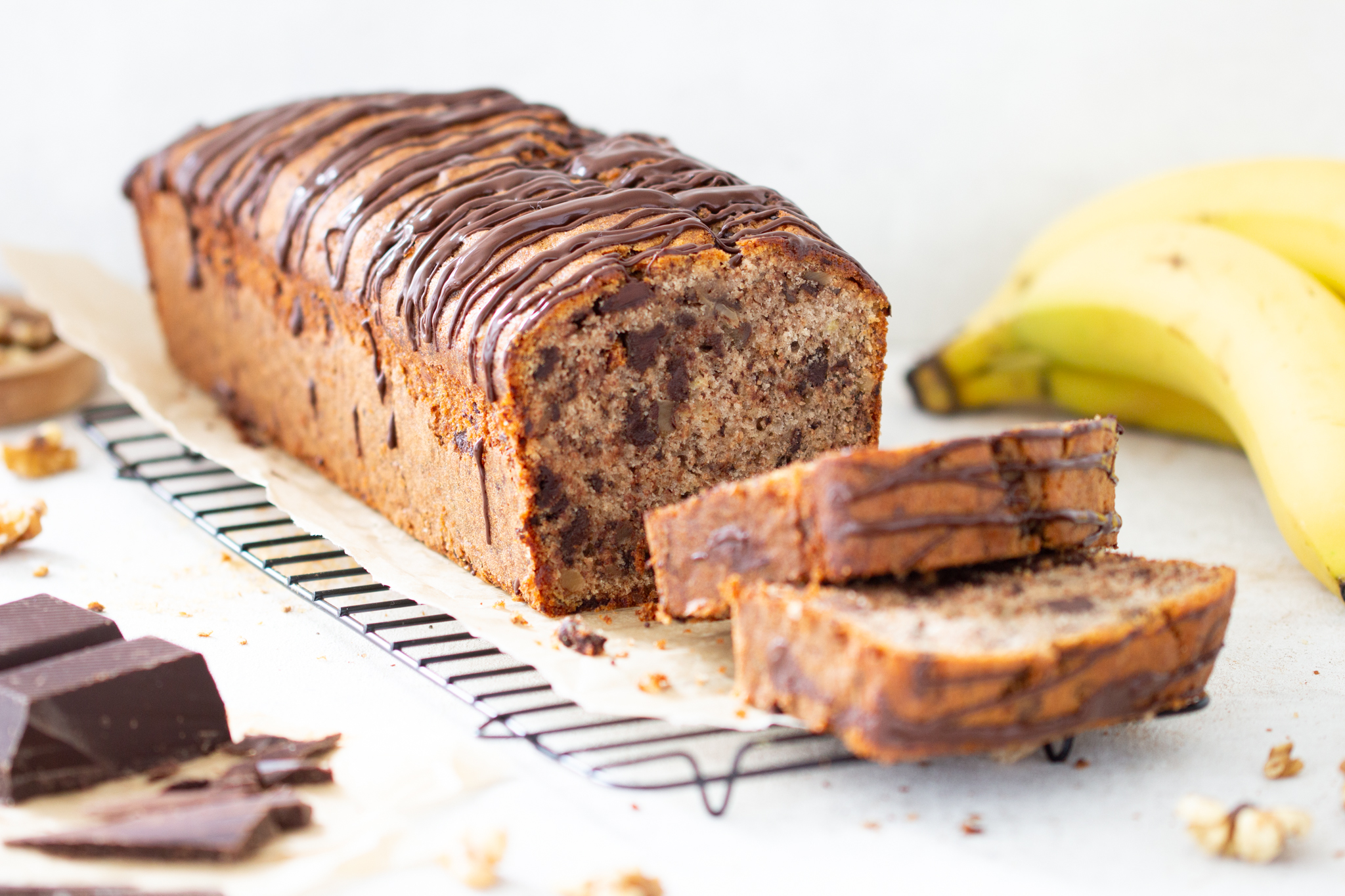 Bananenkuchen aka Bananenbrot als Kastenkuchen mit aufgeschnittenen Stücken. Im Hintergrund liegen Bananen, Walnüsse und ein großes Stück Schokolade